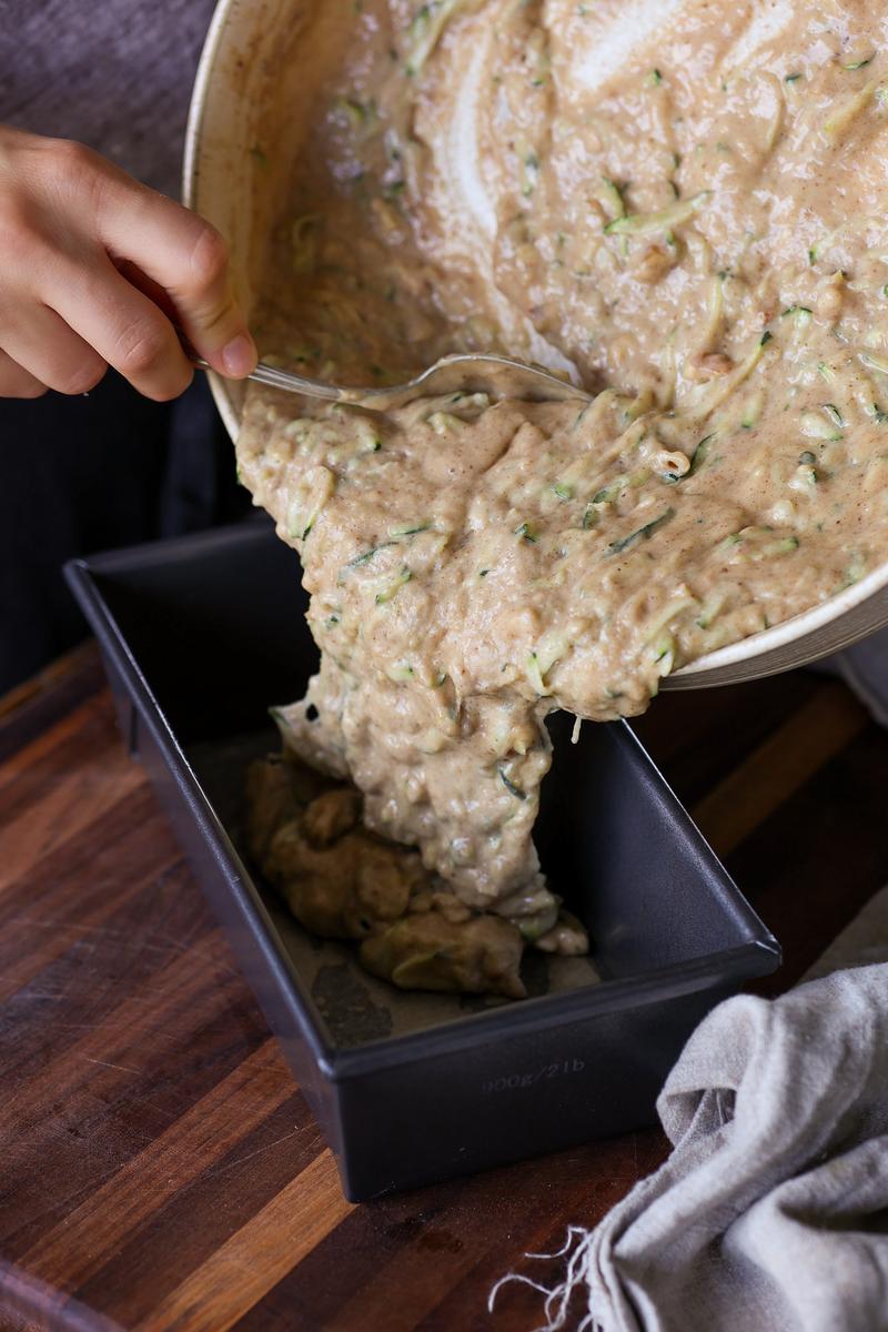 a vegan pouring no egg zucchini bread batter into a loaf pan with pieces of sliced courgette