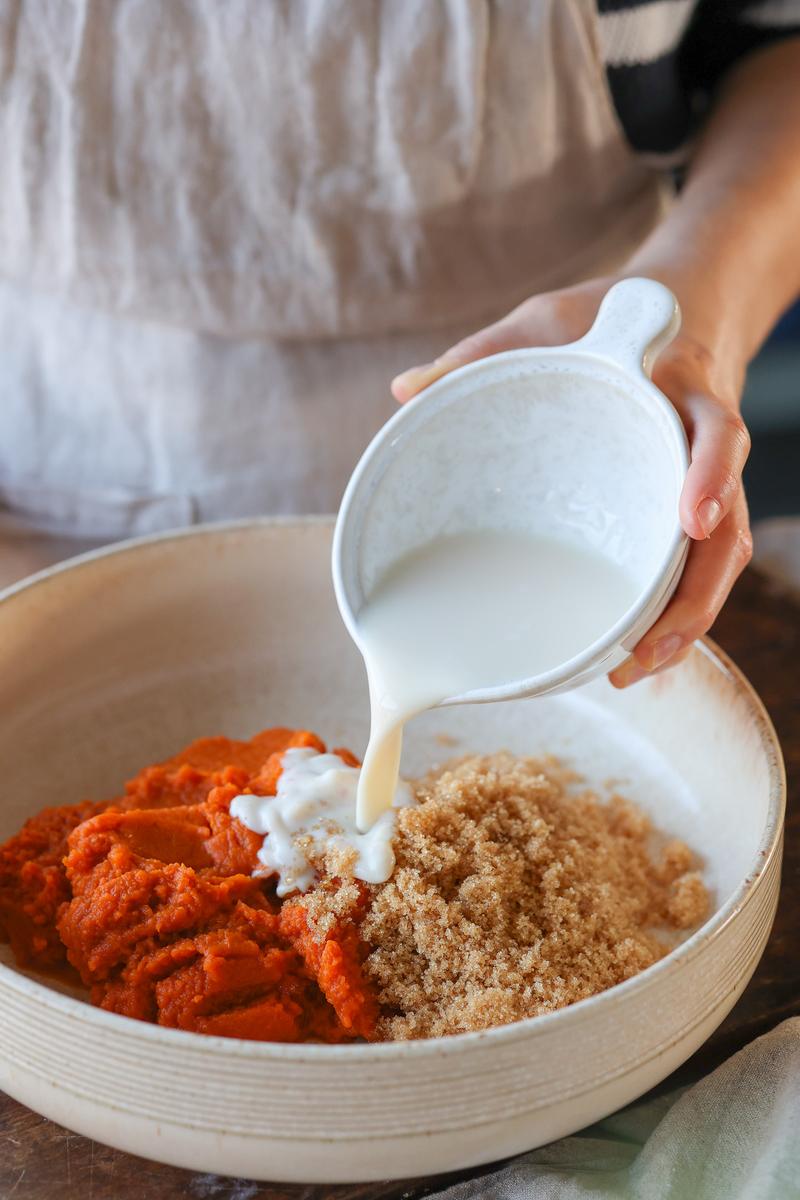 a plant-based person adding soy milk to a large bowl containing pumpkin puree and brown sugar